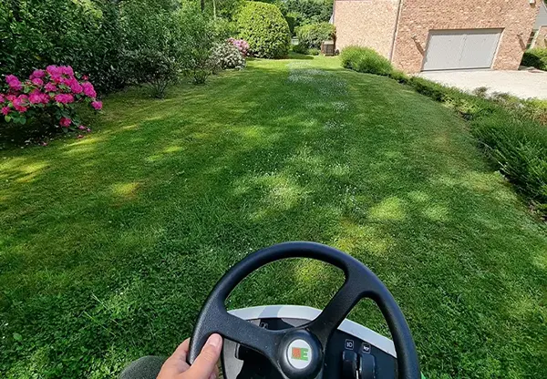 Une personne conduisant une tondeuse sur une pelouse verte, entretenant un jardin fleuri autour d'une belle maison.